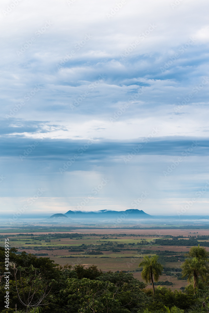 Beautiful view of the hills and open field in Paraguari, Paraguay, with the rain falling over the hills on the horizon and layers of clouds above them, trees in the foreground. Beautiful view of the hills and open field in Paraguari, Paraguay, with the rain falling over the hills on the horizon and layers of clouds above them, trees in the foreground.