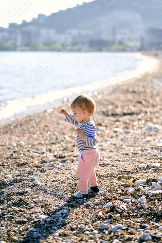 Kid stands on a pebble beach near the sea with his hands up © Nadtochiy