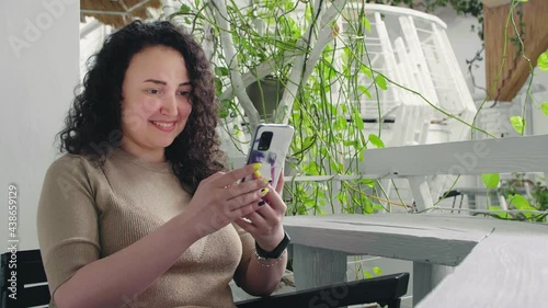 Young cheerful curly girl flips the phone and smiles sitting at the table in the restaurant
