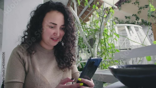 Young cheerful curly girl flips the phone and smiles sitting at the table in the restaurant