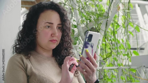 Young serious curly girl flips the phone sitting at the table in the restaurant