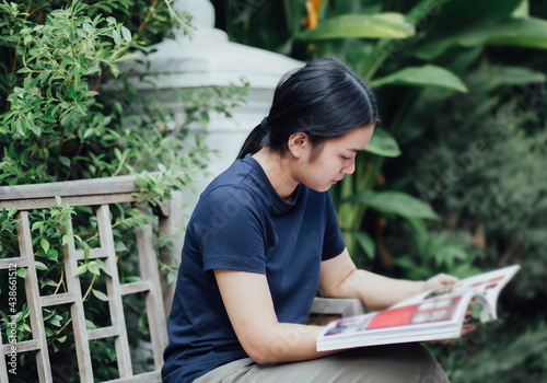 Happy beautiful young Asian girl holds and reading magazines while sitting on a wooden chair in the garden. young woman Relaxing and reading a book in-home yard or the park. a hobby in free time 