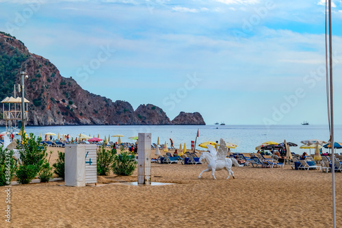 Fototapeta Naklejka Na Ścianę i Meble -  Alanya, Turkey - October 23, 2020: White plastic sculpture of pegasus on Damlatas beach in Alanya. Horse with wings among the yellow sand against the background of sun loungers with umbrellas