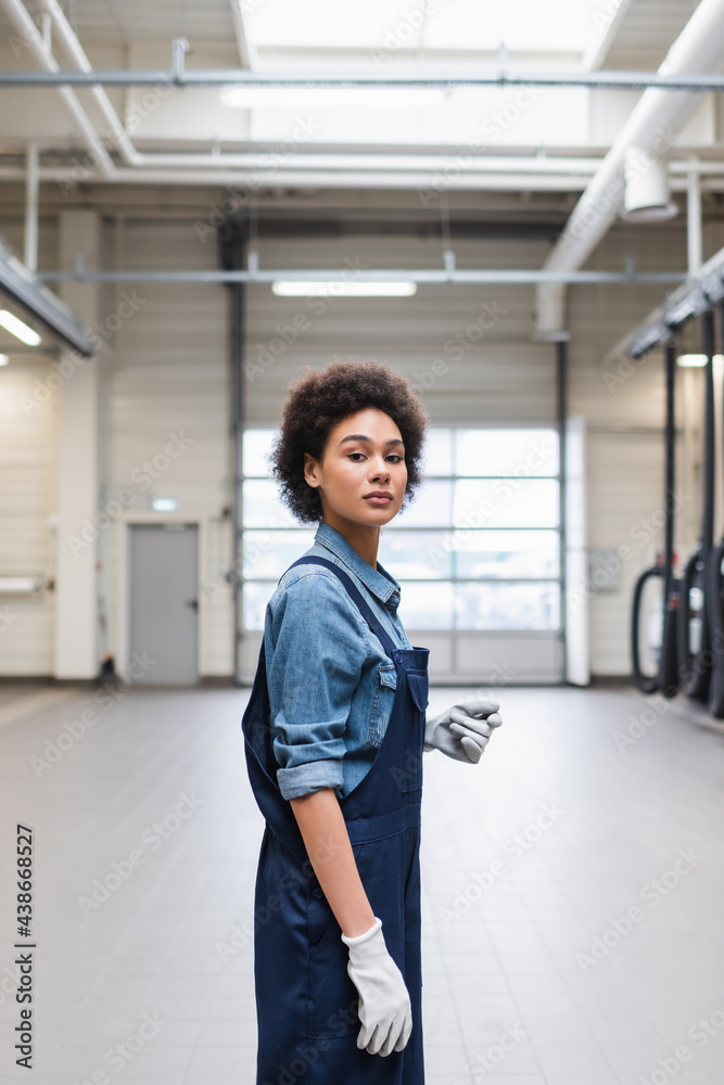 confident young african american mechanic in gloves standing and looking at camera in garage confident young african american mechanic in gloves standing and looking at camera in garage