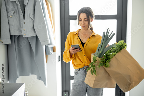 Foto Woman comes home with bags full of fresh groceries, standing with phone at the hallway