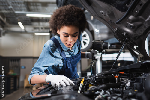 young african american mechanic in overalls holding electric screwdriver and working with car motor in garage photo