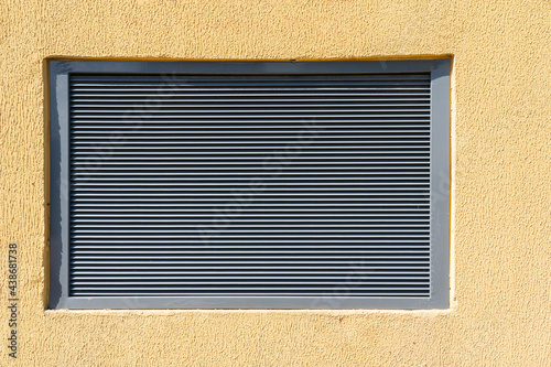 A gray metal window in a yellow stone wall. There is a louvred ventilation grill. Background. Texture.