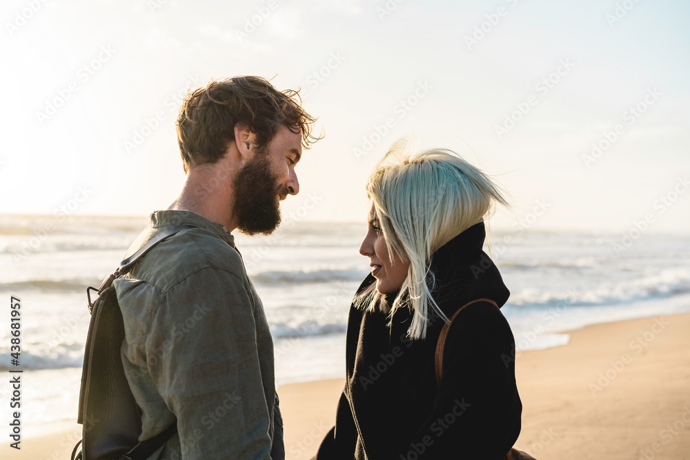 Romantic happy couple in love standing on shoreline at the wild beach - Boyfriend and girlfriend wearing cool clothing and vintage backpack standing in front of the sea while she looking the sunset