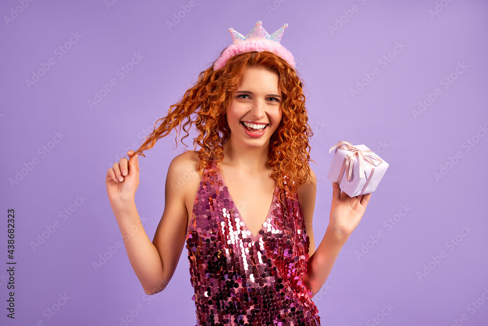Cute red-haired girl princess with curls in a shiny dress and a crown on her head holding a gift isolated on a purple background. The concept of a party, celebration, birthday. Cute red-haired girl princess with curls in a shiny dress and a crown on her head holding a gift isolated on a purple background. The concept of a party, celebration, birthday.