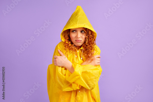 Red-haired sad woman in a yellow raincoat froze, warms herself with her hands isolated on a purple background.