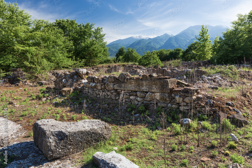 Ruins of an ancient Greek stone building at an archaeological site in northern Greece among trees at the foot of a mountain range on a clear sunny day