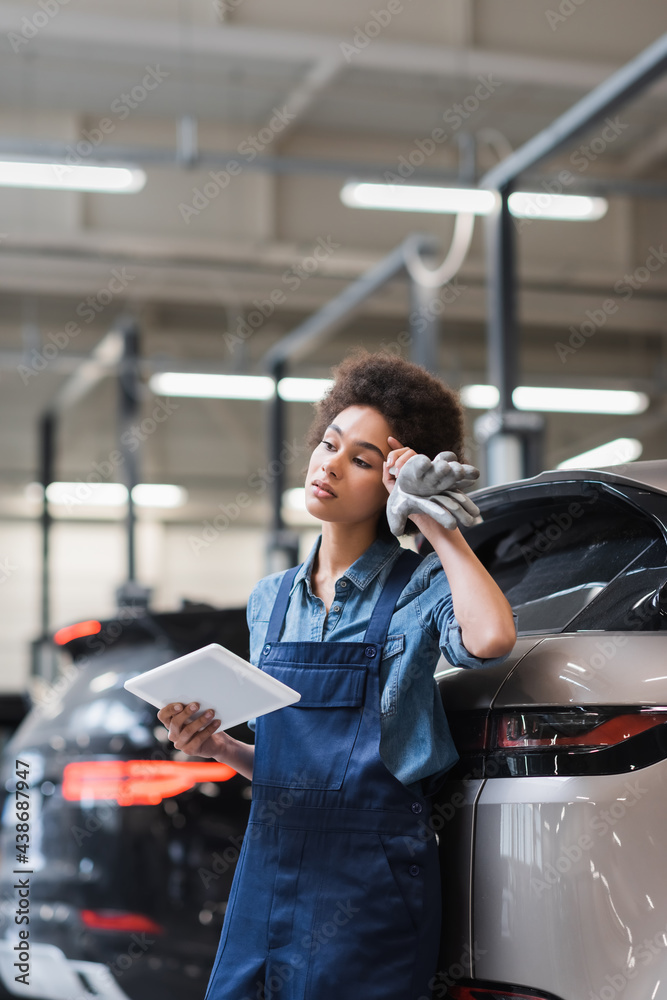 Fototapeta premium tired young african american mechanic standing with digital tablet and holding hand near head in garage