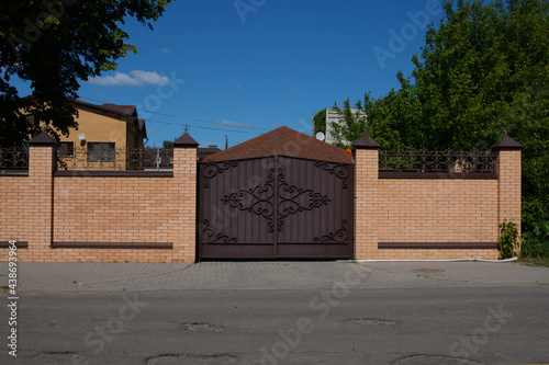 Light brick fence with iron wrought iron gates on the street against a background of wood and blue sky © Sofya