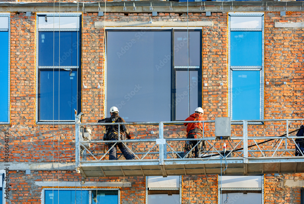 Builders are installing large panoramic windows in a modern residential building using a winch and a mobile platform.