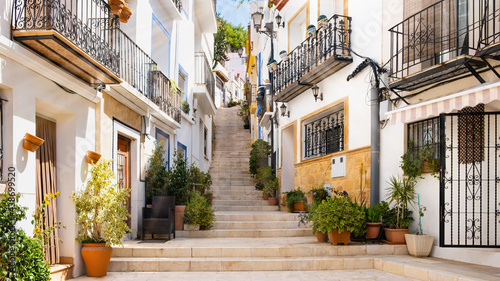 Narrow street with steps, white houses and potted plants in ancient neighborhood El Barrio or Casco Antiguo Santa Cruz in Alicante old town on hillside. Costa Blanca on Mediterranean sea coast, Spain