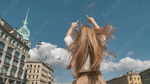Blonde with long loose hair waved by wind poses raising hands against architectural buildings and birds sailing in boat backside photo