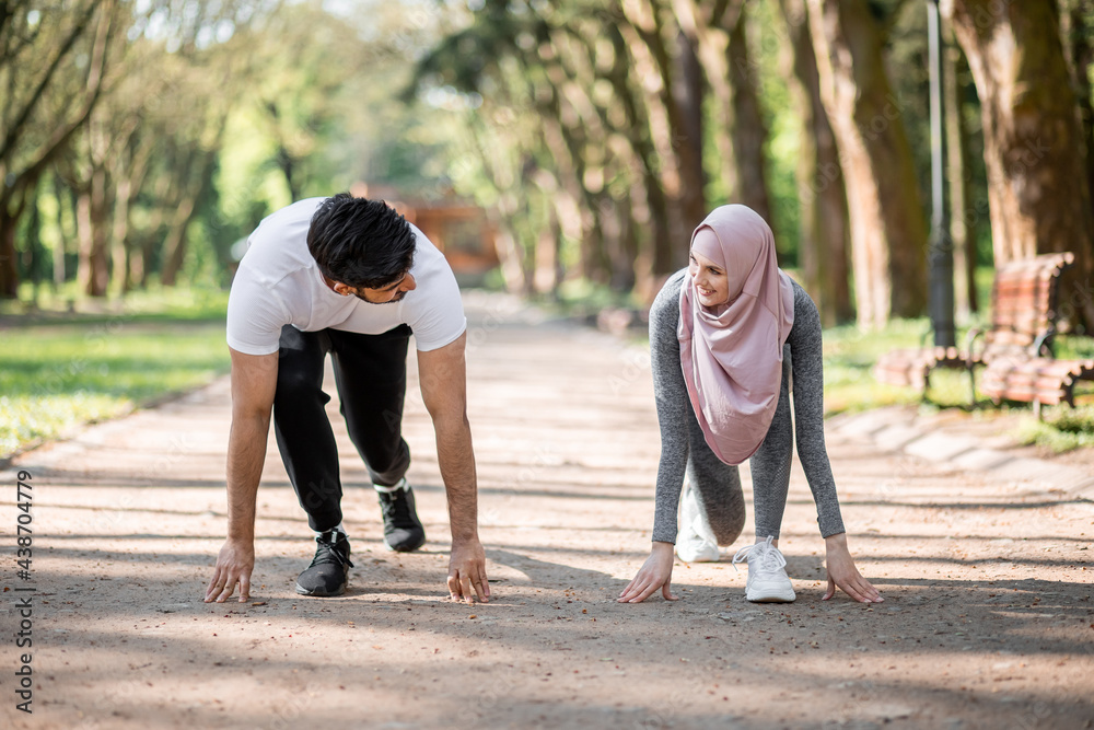 Young muslim couple in sport clothes standing in start position for run at green summer park. Man and woman in hijab preparing for morning workout outdoors. Young muslim couple in sport clothes standing in start position for run at green summer park. Man and woman in hijab preparing for morning workout outdoors.