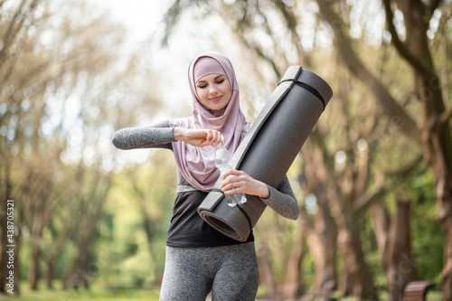 Attractive muslim woman in sport outfit drinking fresh water from bottle after morning exercise at green park. Pretty female in hijab holding yoga mat in hands.