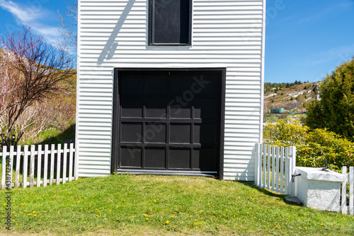 A tall double story white wooden clapboard siding building with a vintage wooden black garage door. There's a small black door above the structure. A white wooden picket fence encloses the garage.