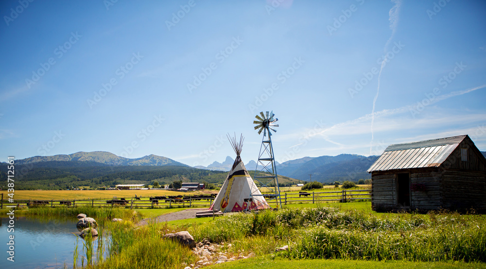 A lake, a teepee and a windmill with the Tetons in the background on a ranch in Alta, Wyoming, on the west side of the Teton Mountains. - obrazy, fototapety, plakaty 