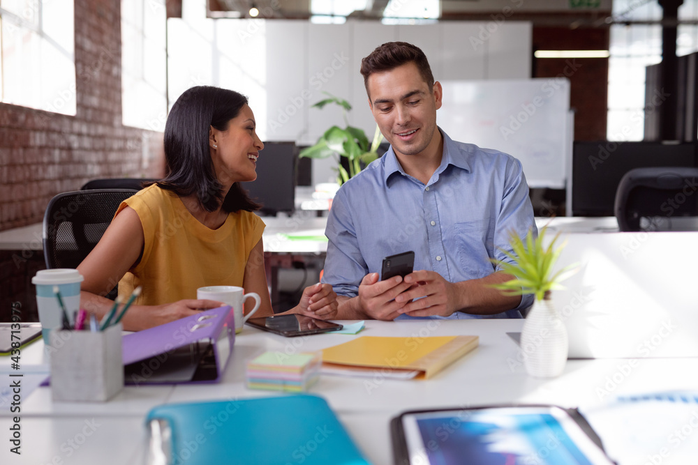 Happy caucasian male and female colleague sitting at desk using smartphone and drinking coffee