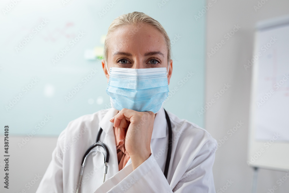 Caucasian female doctor wearing face mask sitting at desk in office listening during video call Caucasian female doctor wearing face mask sitting at desk in office listening during video call