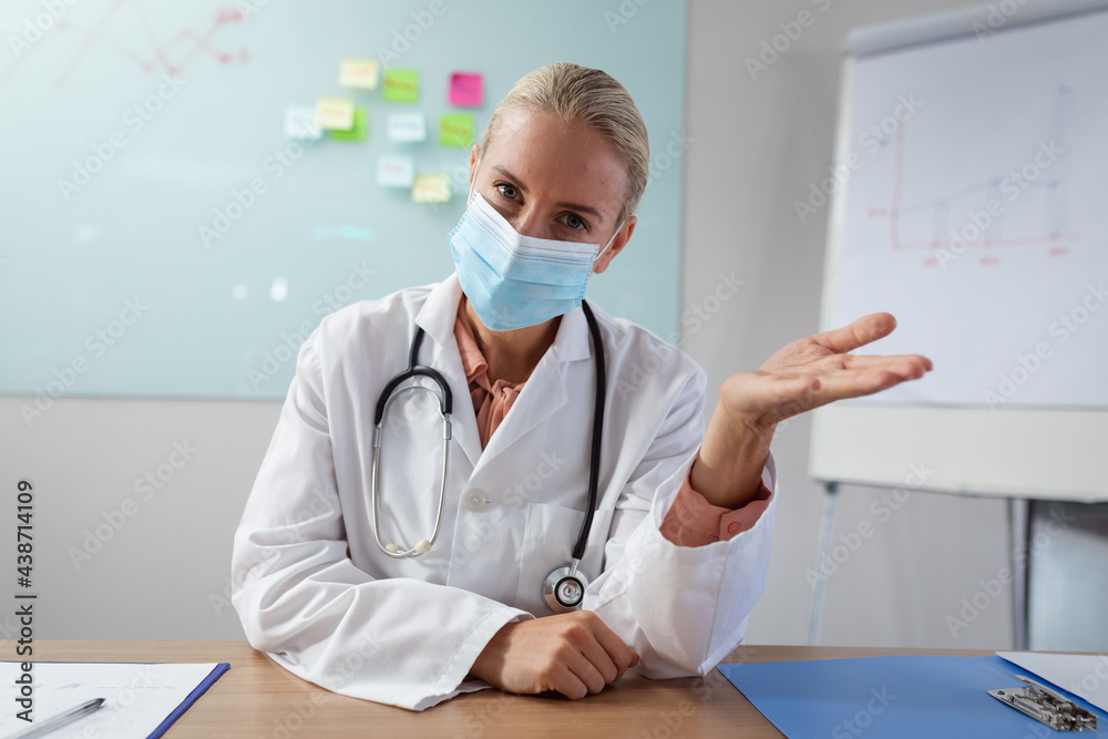 Caucasian female doctor wearing face mask sitting at desk in office gesturing during video call Caucasian female doctor wearing face mask sitting at desk in office gesturing during video call