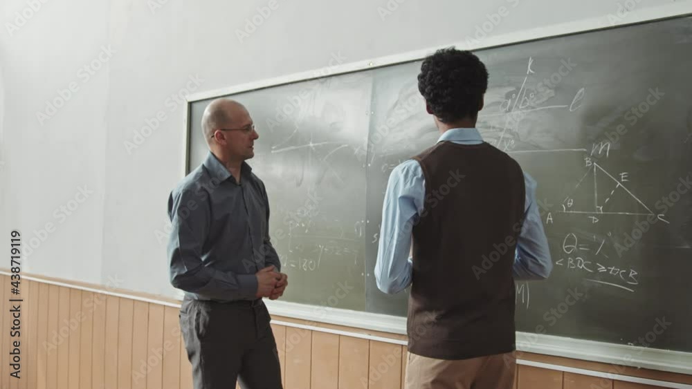 Aerial medium slowmo shot of male Mixed-race student answering at blackboard trying to solve hard Geometry problem with help of mid-adult professor
