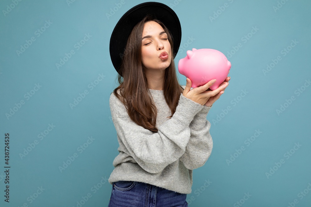 Portrait photo of happy positive young beautiful attractive brunette woman with sincere emotions wearing grey sweater and black hat isolated over blue background with copy space and holding pink piggy Portrait photo of happy positive young beautiful attractive brunette woman with sincere emotions wearing grey sweater and black hat isolated over blue background with copy space and holding pink piggy