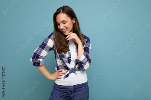 Photo portrait of young beautiful smiling hipster brunette woman in trendy blue and white shirt and jeans clothes. Sexy carefree female person posing isolated near blue wall with empty space in studio © Ivan Traimak