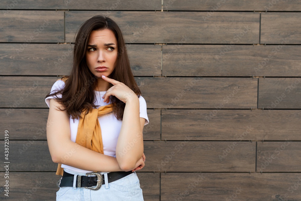 Fototapeta premium Portrait of touchy sad upset offended resentful young brunet woman wearing casual white t-shirt and jeans with yellow sweater poising near brown wall in the street