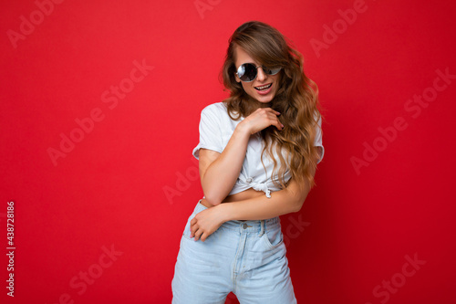 Photo shot of beautiful sexy positive young blonde woman wearing casual clothes and stylish sunglasses isolated over colorful background looking at camera © Ivan Traimak