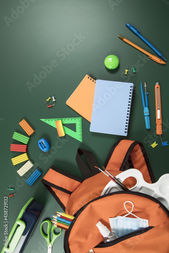Office supplies in an orange school backpack lie on a green background. School supplies lie on the green chalkboard. Vertical, lot of free space for text. Education concept.
