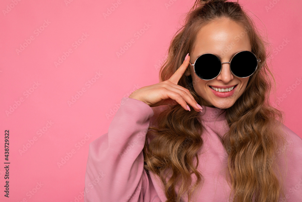 Closeup photo of pretty positive smiling young blonde curly woman isolated over pink background wall wearing casual pink sport clothes and stylish sunglasses looking at camera Closeup photo of pretty positive smiling young blonde curly woman isolated over pink background wall wearing casual pink sport clothes and stylish sunglasses looking at camera