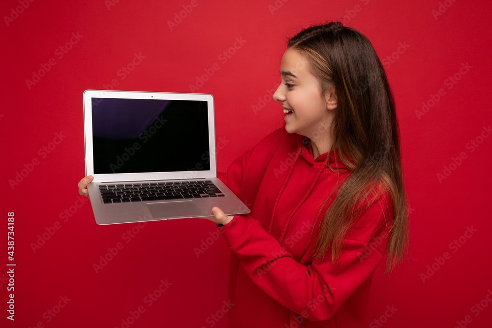 Side profile photo of beautiful happy girl with long hair wearing red hoodie holding computer laptop looking at netbook keyboard and screen monitor isolated over red wall Side profile photo of beautiful happy girl with long hair wearing red hoodie holding computer laptop looking at netbook keyboard and screen monitor isolated over red wall