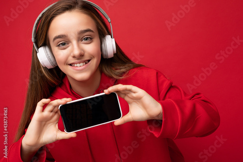 Closeup portrait of amazibg beautiful brunette girl wearing red hoodie isolated on red background holding and showing smartphone with empty display for cutout wearing white wireless headsets listening photo