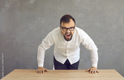 Billede på lærred Studio portrait of an angry stressed young man in glasses leaning over his desk and shouting