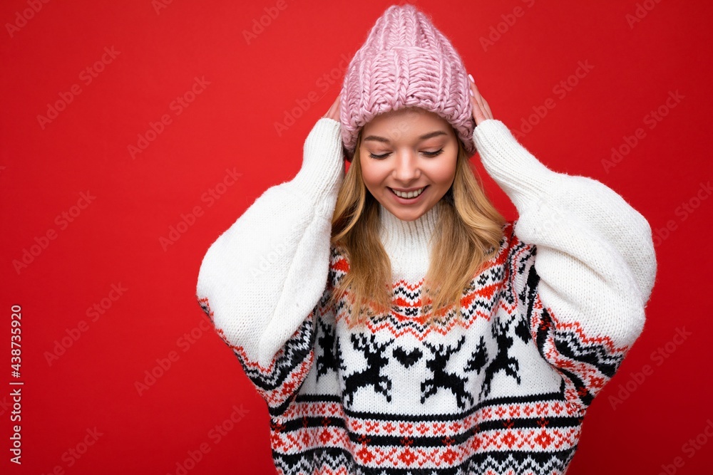 Photo of beautiful happy cute young blonde woman isolated over red background wall wearing winter sweater and pink hat looking down Photo of beautiful happy cute young blonde woman isolated over red background wall wearing winter sweater and pink hat looking down