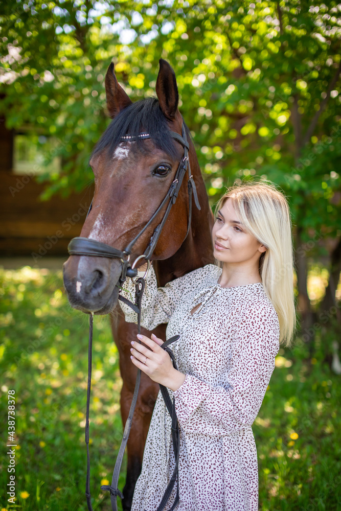 Obraz premium A young rider woman blonde with long hair in a dress posing with brown horse in forest next to farm