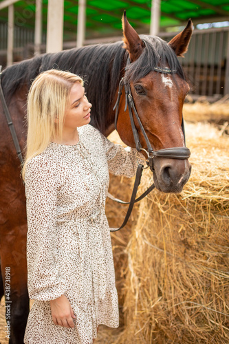 A young rider woman blonde with long hair in a dress posing with brown horse inside light stable, Russia
