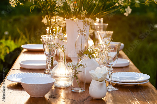 Elegant wedding dinner table decorated with white dishes, flowers in glass vases. Festive atmosphere, luxury party. Sunset, bright summer evening. Close up, details