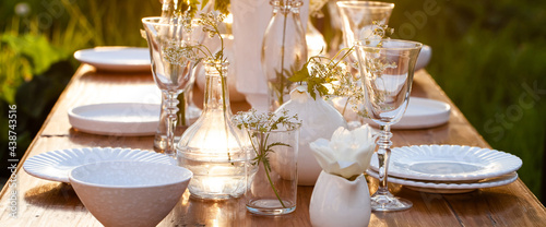 Elegant wedding dinner table decorated with white dishes, flowers in glass vases. Festive atmosphere, luxury party. Sunset, bright summer evening. Close up, details. Banner photo
