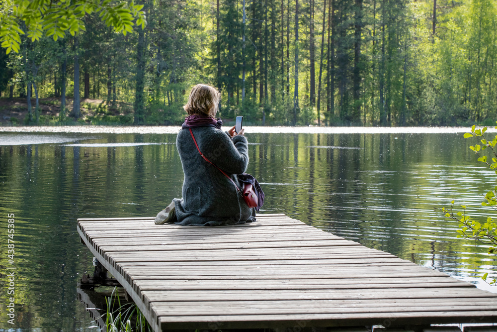 A young woman takes photos with a smartphone sitting on the wooden pier on a forest lake on a sunny summer day. A young woman takes photos with a smartphone sitting on the wooden pier on a forest lake on a sunny summer day.