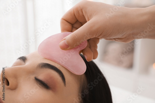 Young woman receiving facial massage with gua sha tool in beauty salon, closeup photo