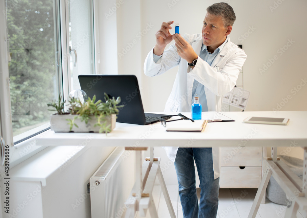 male chemist, scientist works at standing table and examines blue liquids in small vials and wears white coat
