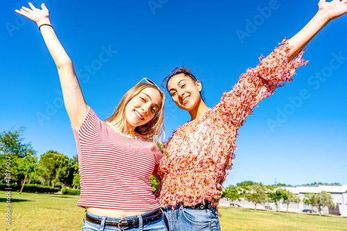 Two happy girls embracing each other with open up arms smiling looking at camera in a city park field in sunny day with blue sky. Mixed young women couple enjoying nature celebrating their diversity