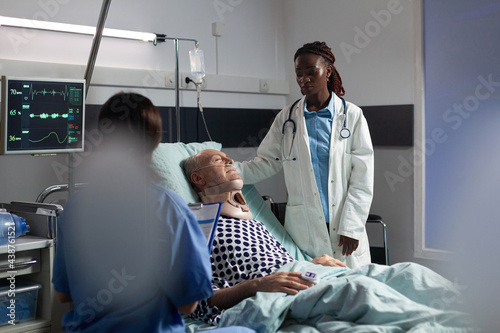 Injured senior man with neck brace laying in bed suffering after accident, discussing with african doctor during medical visit and assistant taking notes on clipboard. about body trauma.