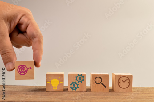 Hand holding dartboard icon with bulb, teamwork, magnifying glass and clock icons on wooden blocks. Business target concept.