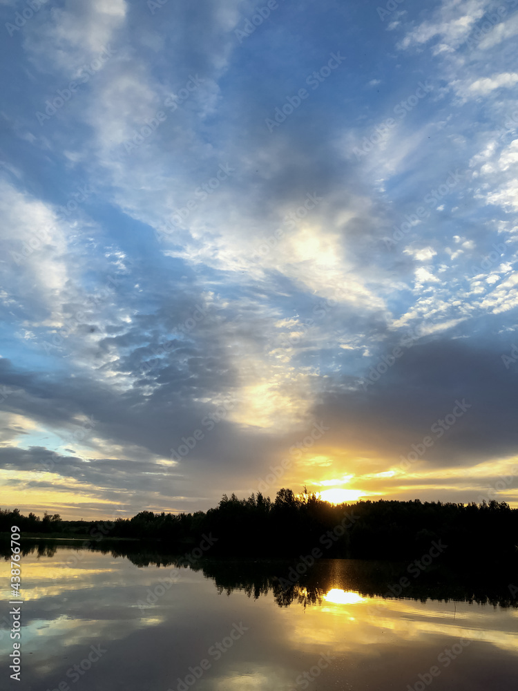 Sunset over the lake. The sun sets behind silhouetted forest on the horizon. Dramatic overcast sky reflects on lake water surface Sunset over the lake. The sun sets behind silhouetted forest on the horizon. Dramatic overcast sky reflects on lake water surface