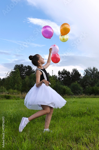 cute girl in beautiful dress is holding colorful balloons in summer meadow against background of blue sky and clouds, having fun and playing. Сoncept of festive mood, birthday